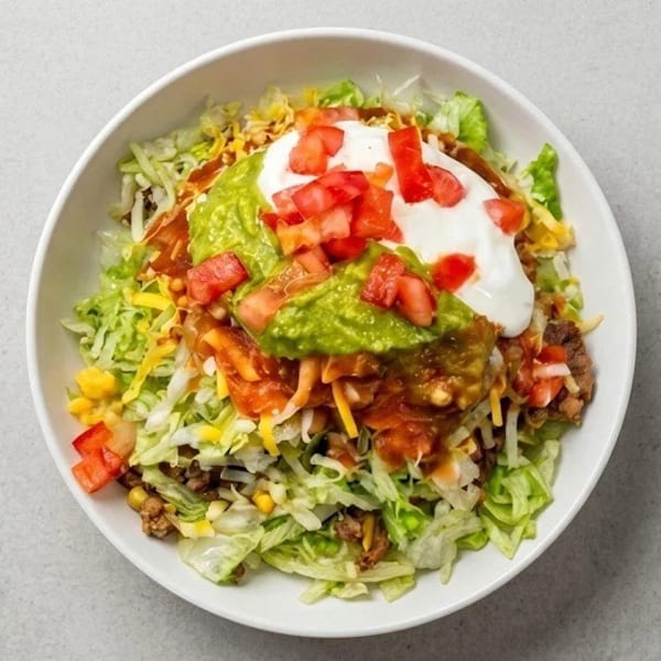 A vibrant taco salad in a white bowl, topped with guacamole, sour cream, and diced tomatoes.