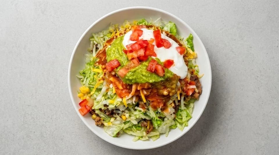 A vibrant taco salad in a white bowl, topped with guacamole, sour cream, and diced tomatoes.