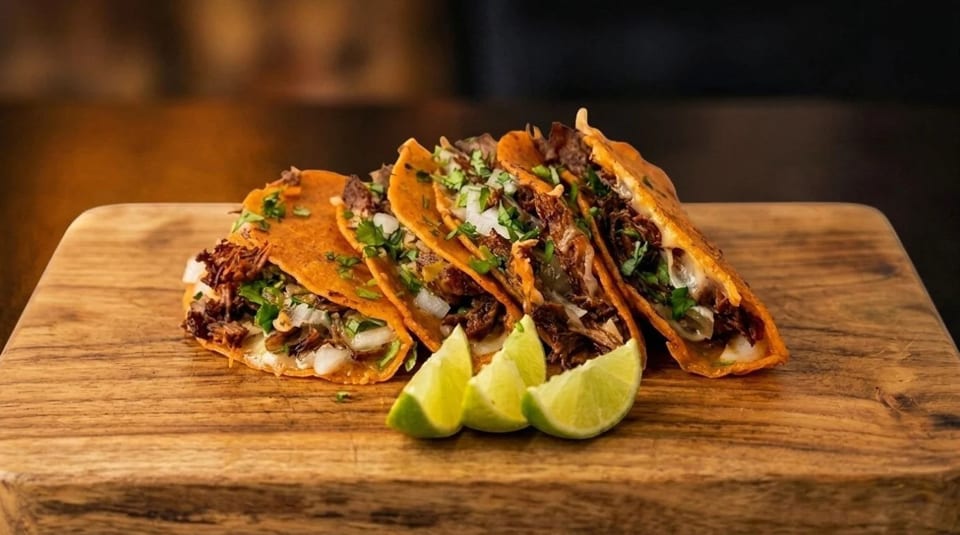 Five birria tacos with cilantro, onions, and lime wedges on a wooden board.